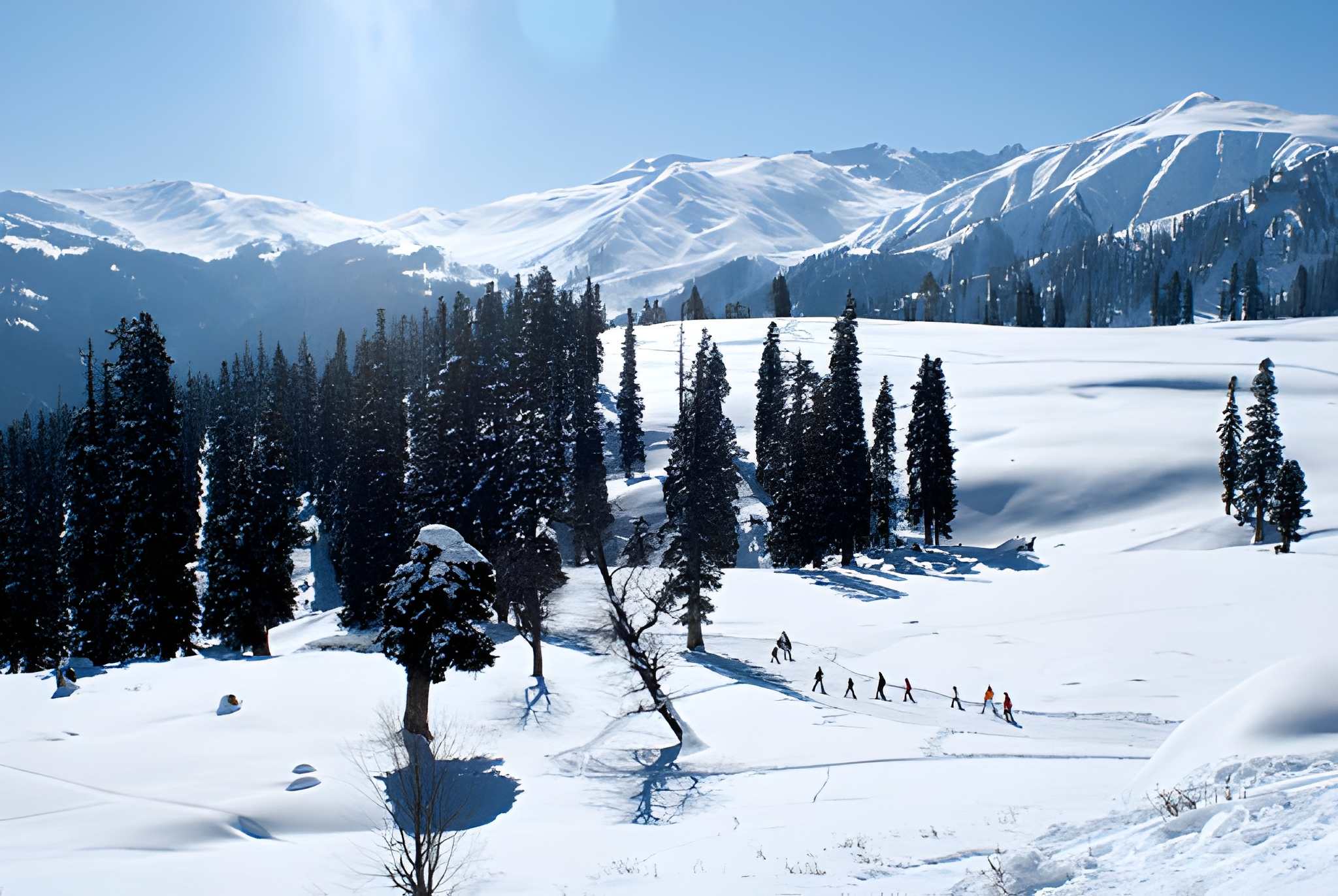 Pine forest and snow-covered Himalayan slopes with a trail of people trekking under a bright blue sky among the Best places to see snow in India for winter adventure lovers.
