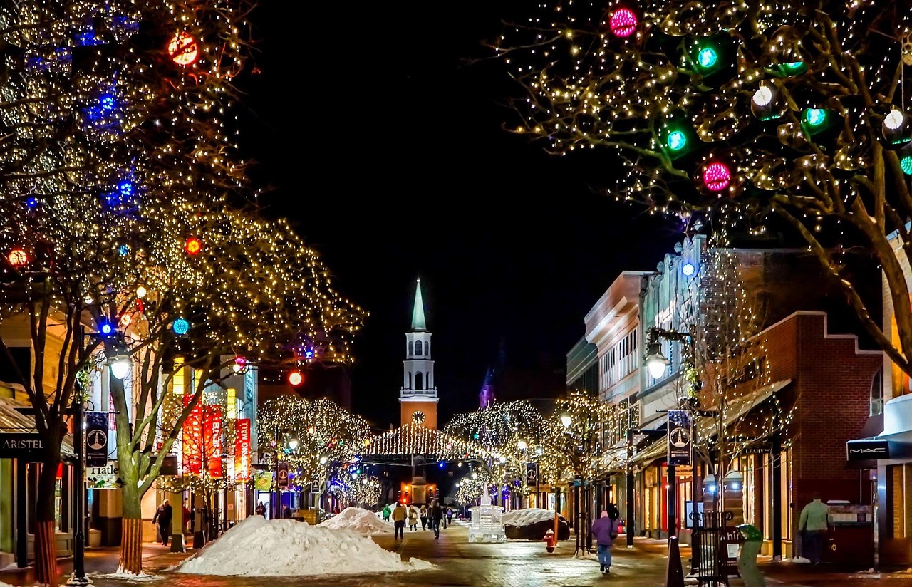 Festive nighttime street with trees covered in warm white and colorful holiday lights, snow piles on a wet cobblestone road, and people walking nearby, reflecting festivals in December.