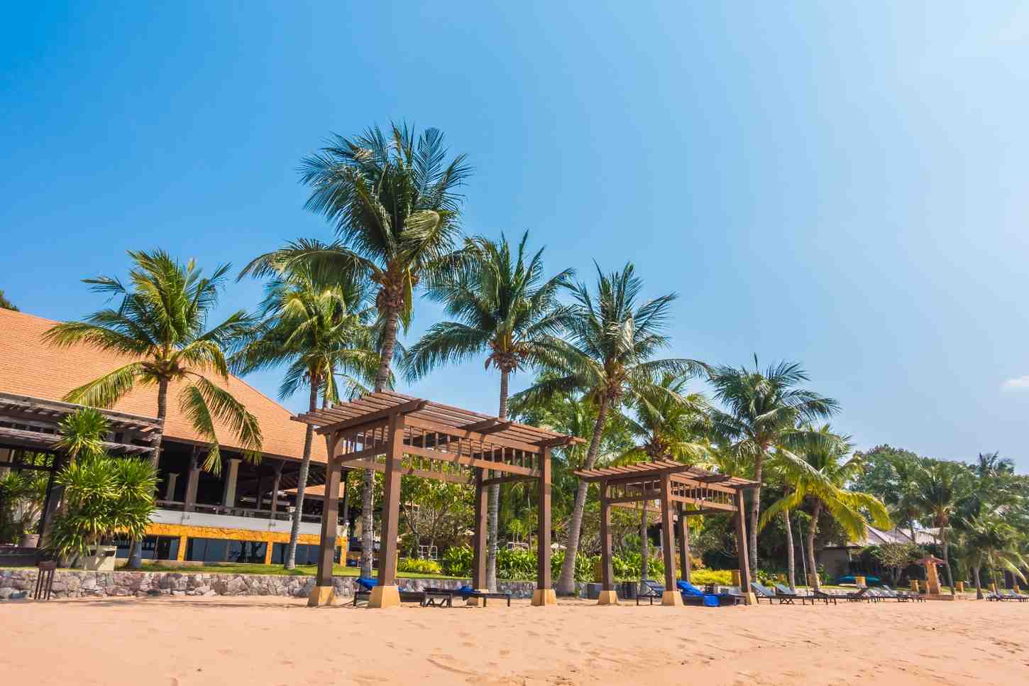 A sunny beachfront lined with palm trees, wooden pergolas, and lounge chairs in front of a resort, offering a calm and refreshing seaside atmosphere among the Things to do in Panjim.