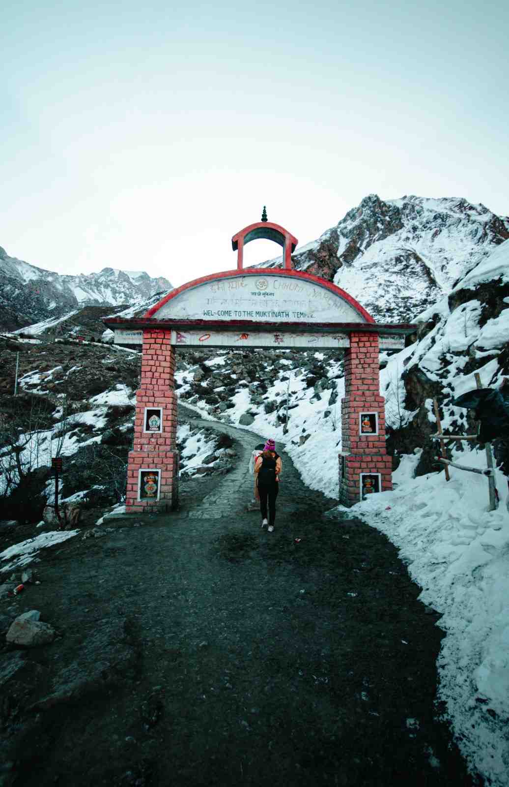 Snow-covered mountain trail leading to the Muktinath Temple entrance gate, with a trekker walking on the path surrounded by winter landscapes—an iconic view often associated with Winter Treks in India.