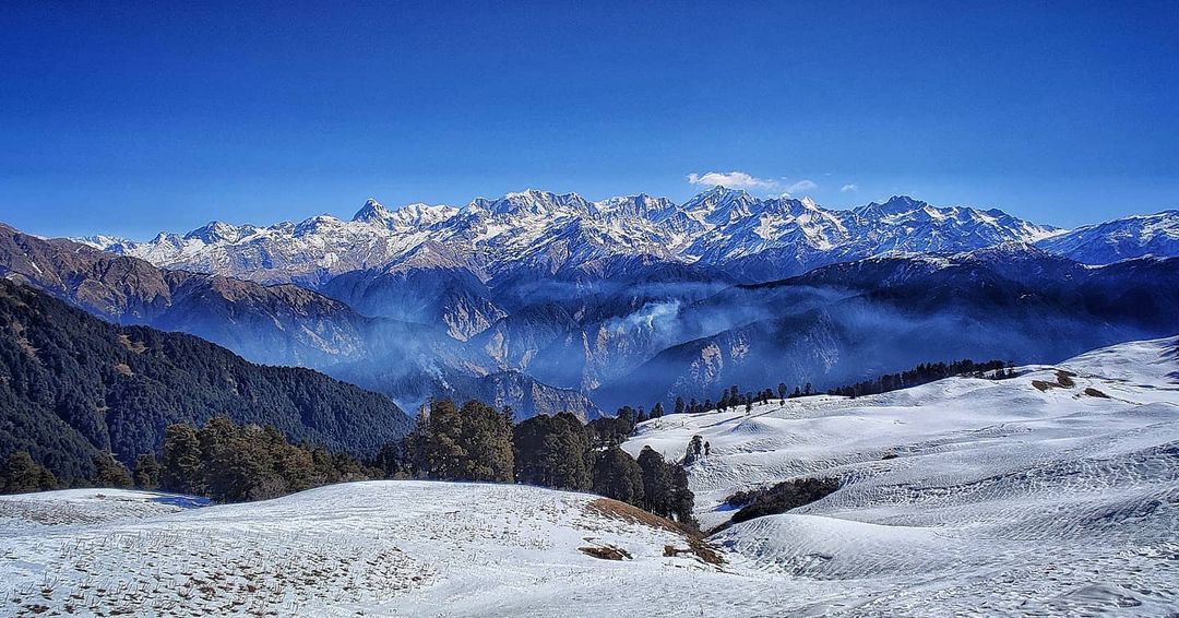 Snow-covered meadows and dense forested ridges with majestic Himalayan peaks glowing under a clear blue sky, showcasing the serene beauty of the best winter treks.