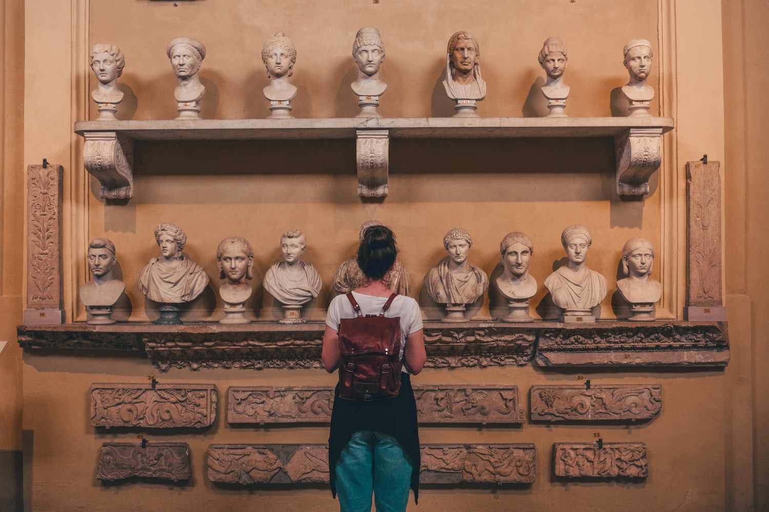 A visitor with a small backpack stands in front of a wall display of ancient marble busts in a softly lit gallery, observing the sculptures arranged on shelves; a scene reminiscent of the quiet, reflective atmosphere often found in Museums in Goa.