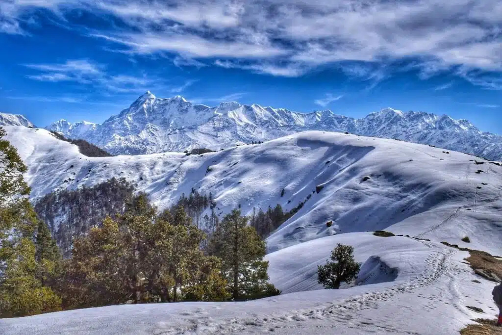 Snow-covered Himalayan landscape with rolling white slopes, pine trees, and majestic high peaks under a blue sky capturing the serene beauty often experienced during Winter Treks in India.
