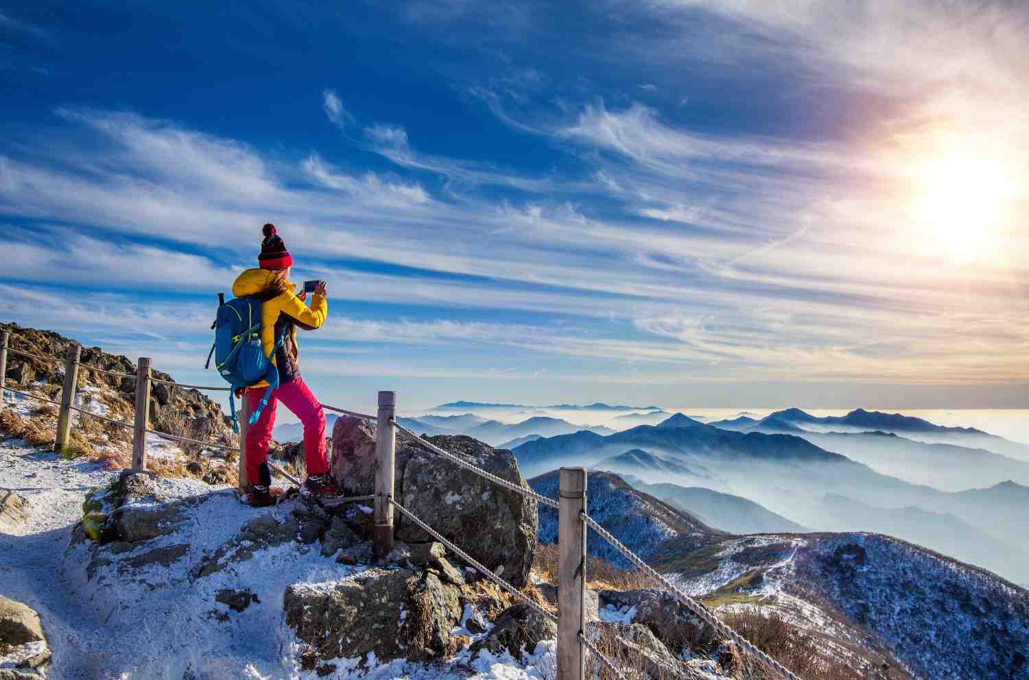 Snow-covered mountain viewpoint where a trekker in bright winter gear photographs the stunning sunrise over misty mountain ranges, showcasing the beauty of the best winter treks.