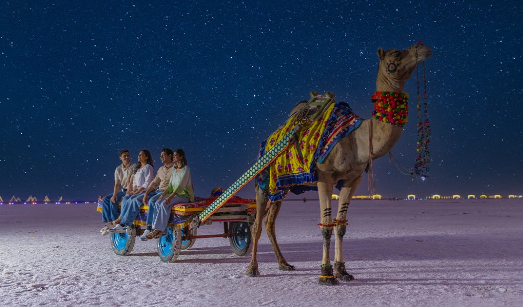 People riding a decorated camel cart at night under a starry sky in a desert setting, representing a december festival.