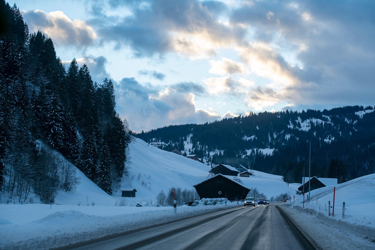 Winter highway passing through snow-covered hills and pine forests, vehicles driving on a slightly wet open road, and dramatic cloudy sky at dusk perfect inspiration for winter road trips.