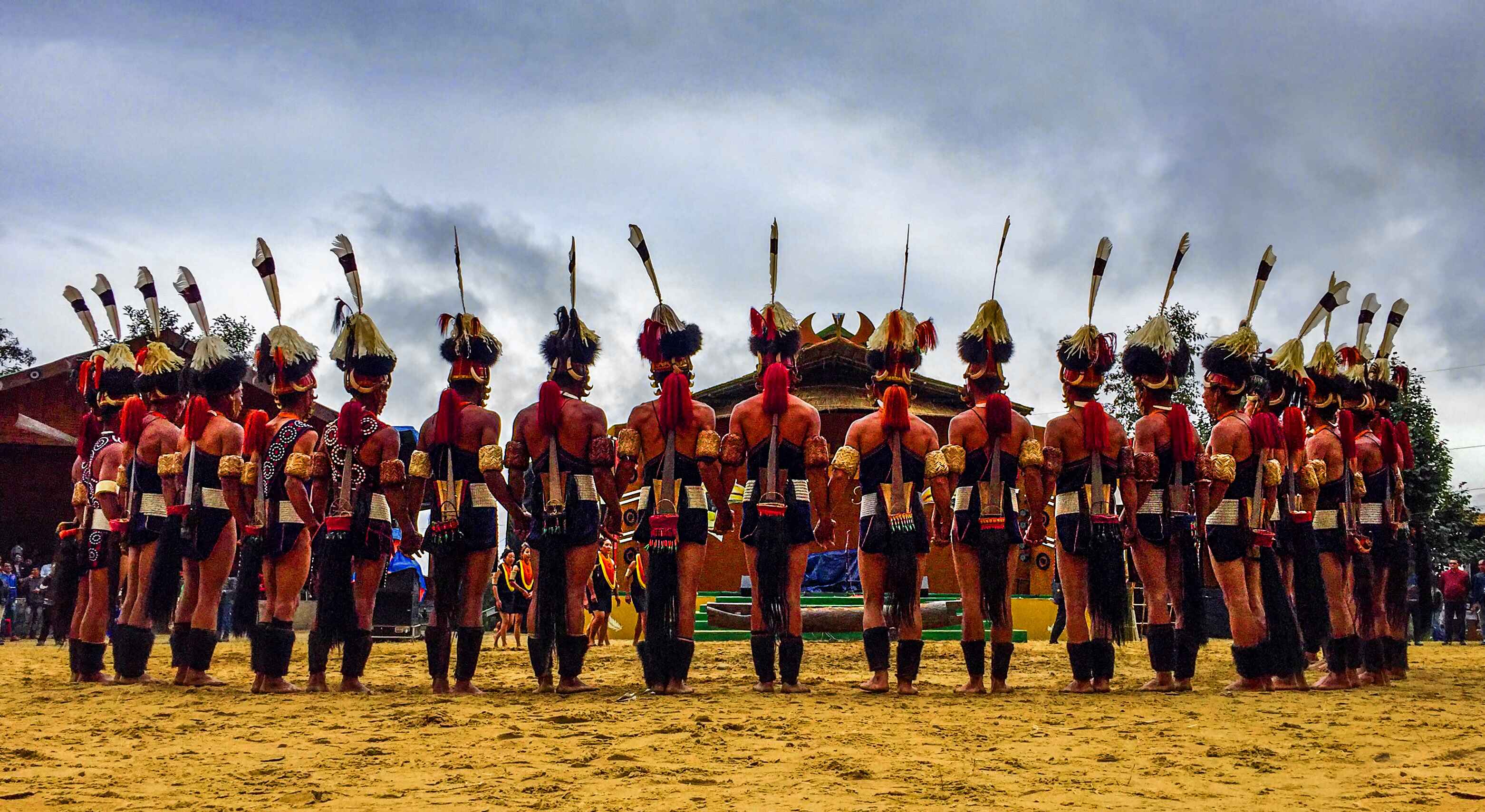 Traditional tribal dancers standing in a formation on a sandy ground wearing feathered headgear and cultural attire under a cloudy sky, showing a celebration style of a December event.
