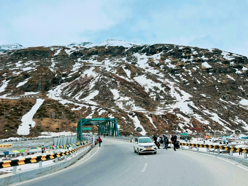 Curved winter mountain road leading to a green steel bridge with snow-patched hills and travelers walking along the roadside an inspiring view for winter road trips in India during December.