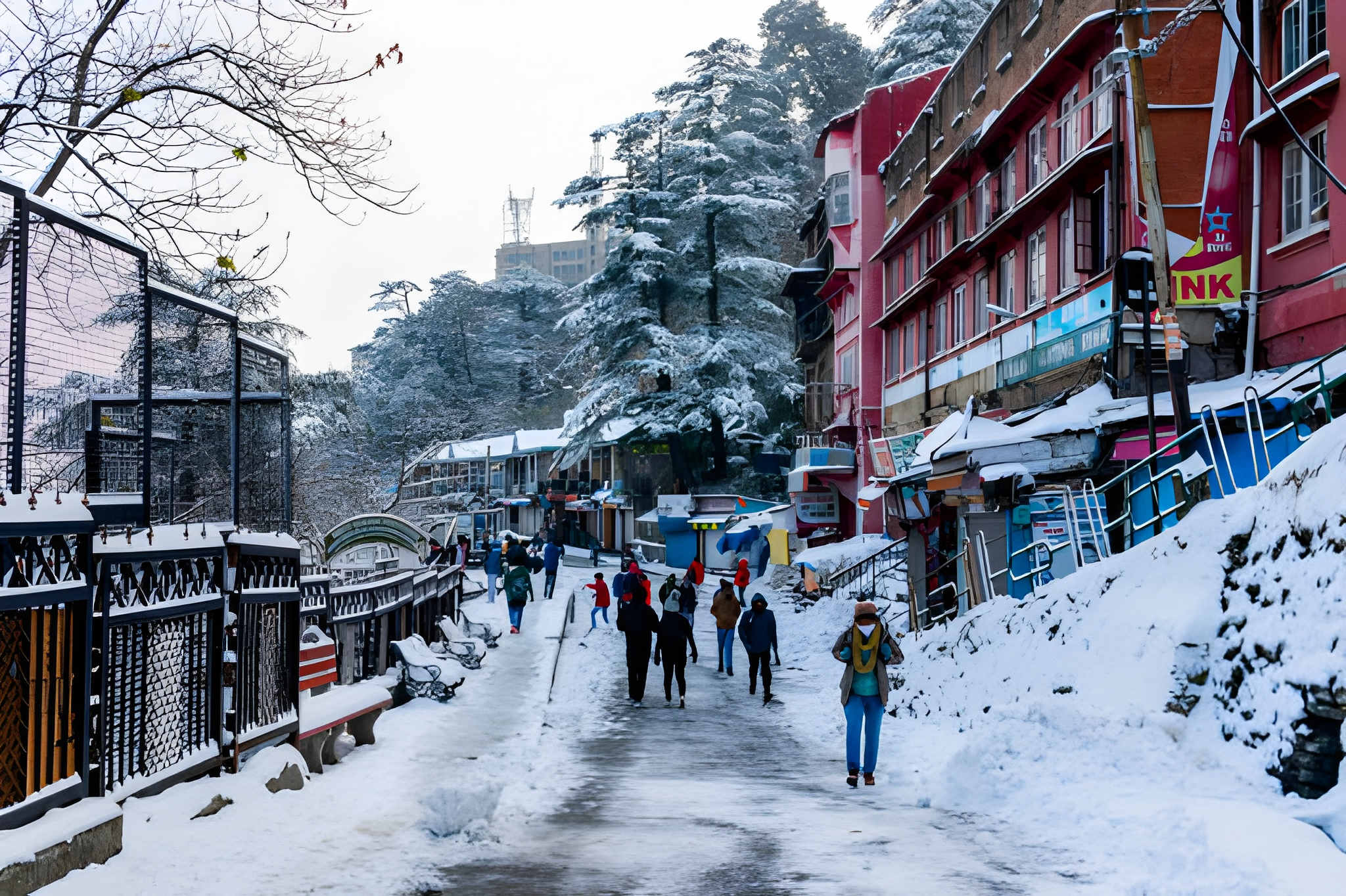 Busy Himalayan hill-town street with snow-covered roofs, trees, and roadside slopes, people walking on a partially wet winter road one of the charming Winter travel destinations in India and a beautiful glimpse of Top Snowfall Destinations.