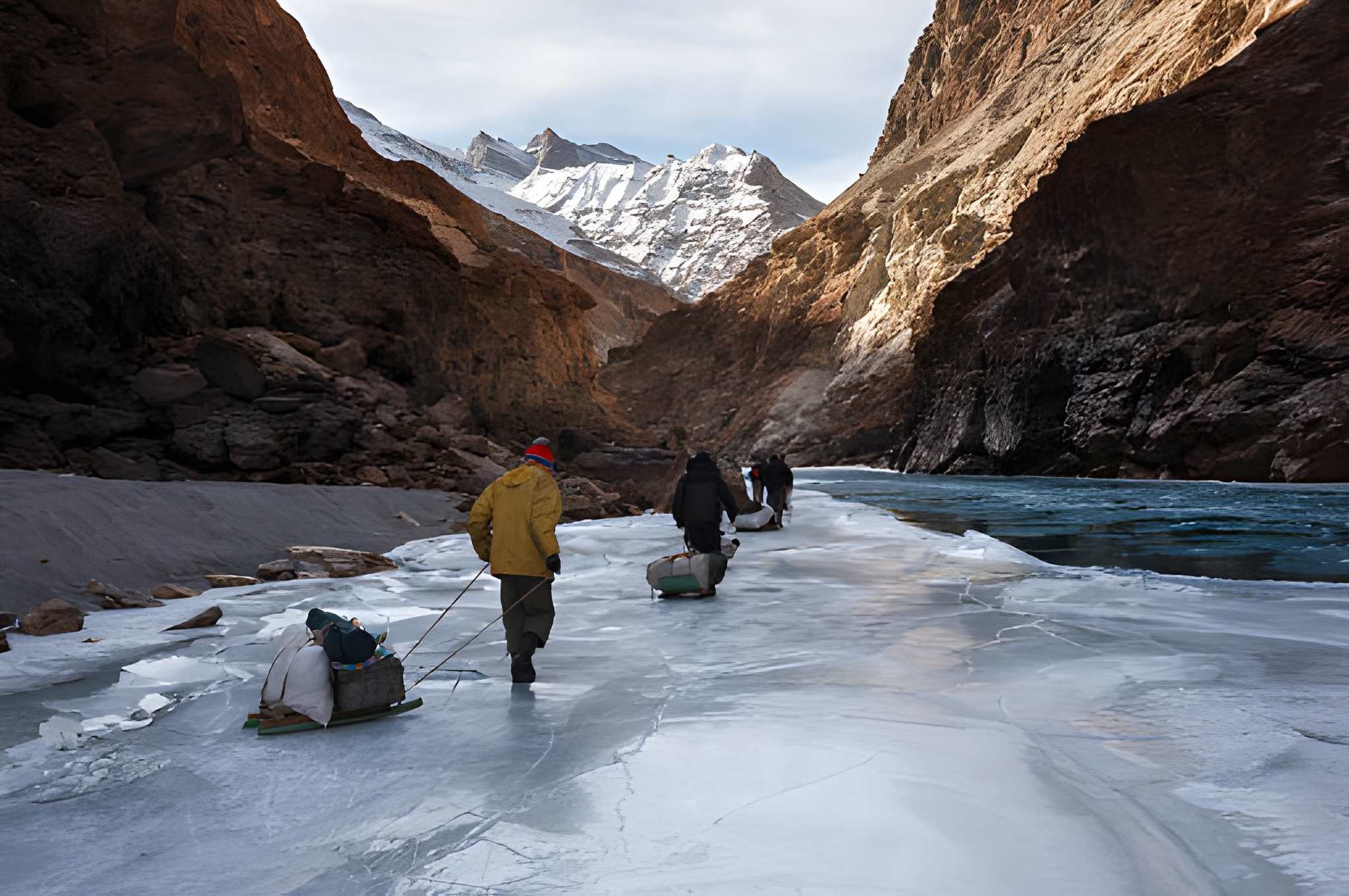 Travelers pulling heavy sleds across a partially frozen river between steep rocky Himalayan cliffs, with snow-capped peaks towering ahead an authentic winter scene from one of the most adventurous Top Snowfall Destinations and a striking moment from India’s Winter travel destinations in India.