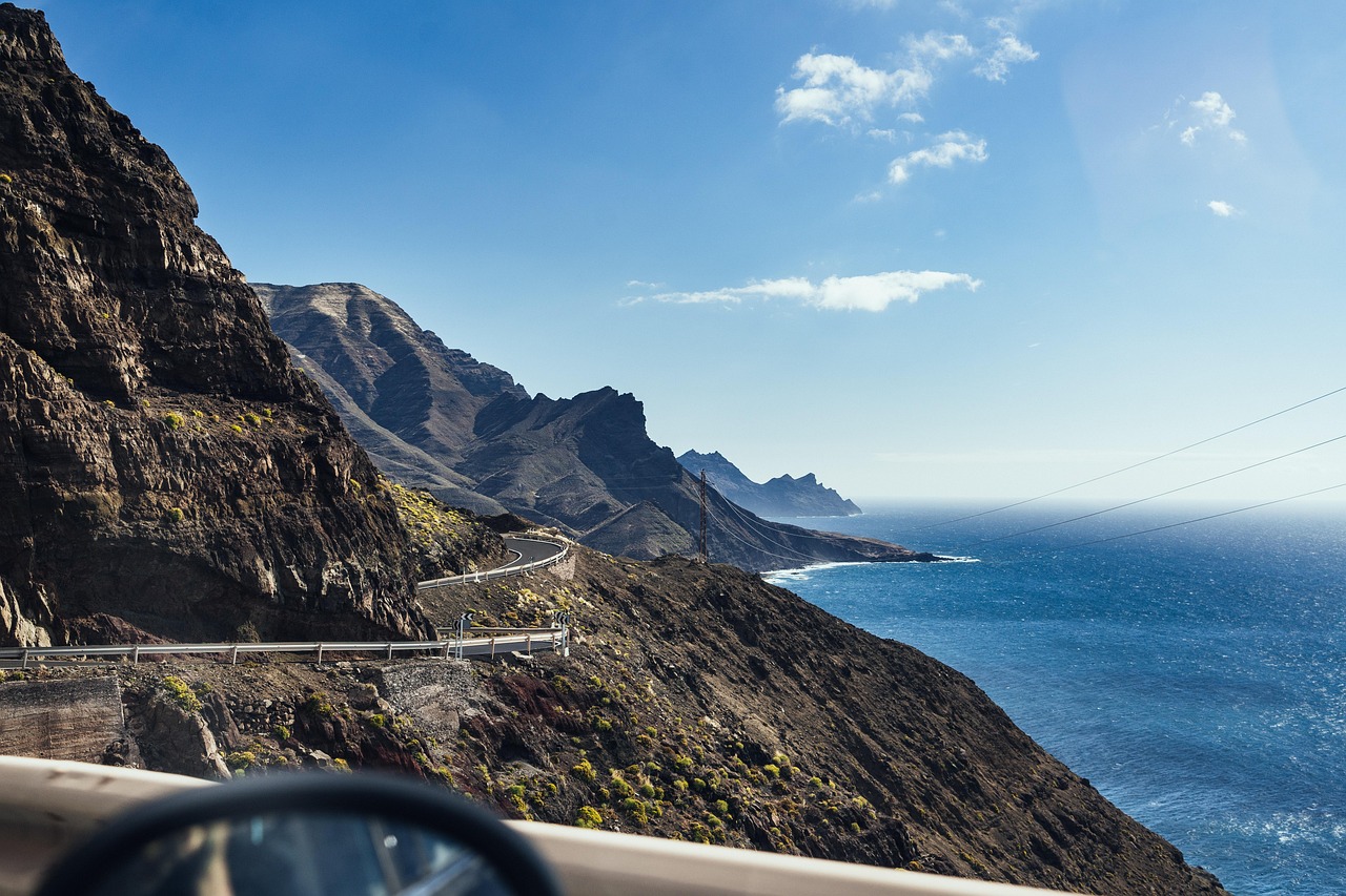 Mountain cliffside road winding along a rugged coastline with the blue ocean stretching into the horizon, rocky peaks on one side, and sunlight reflecting over the water.