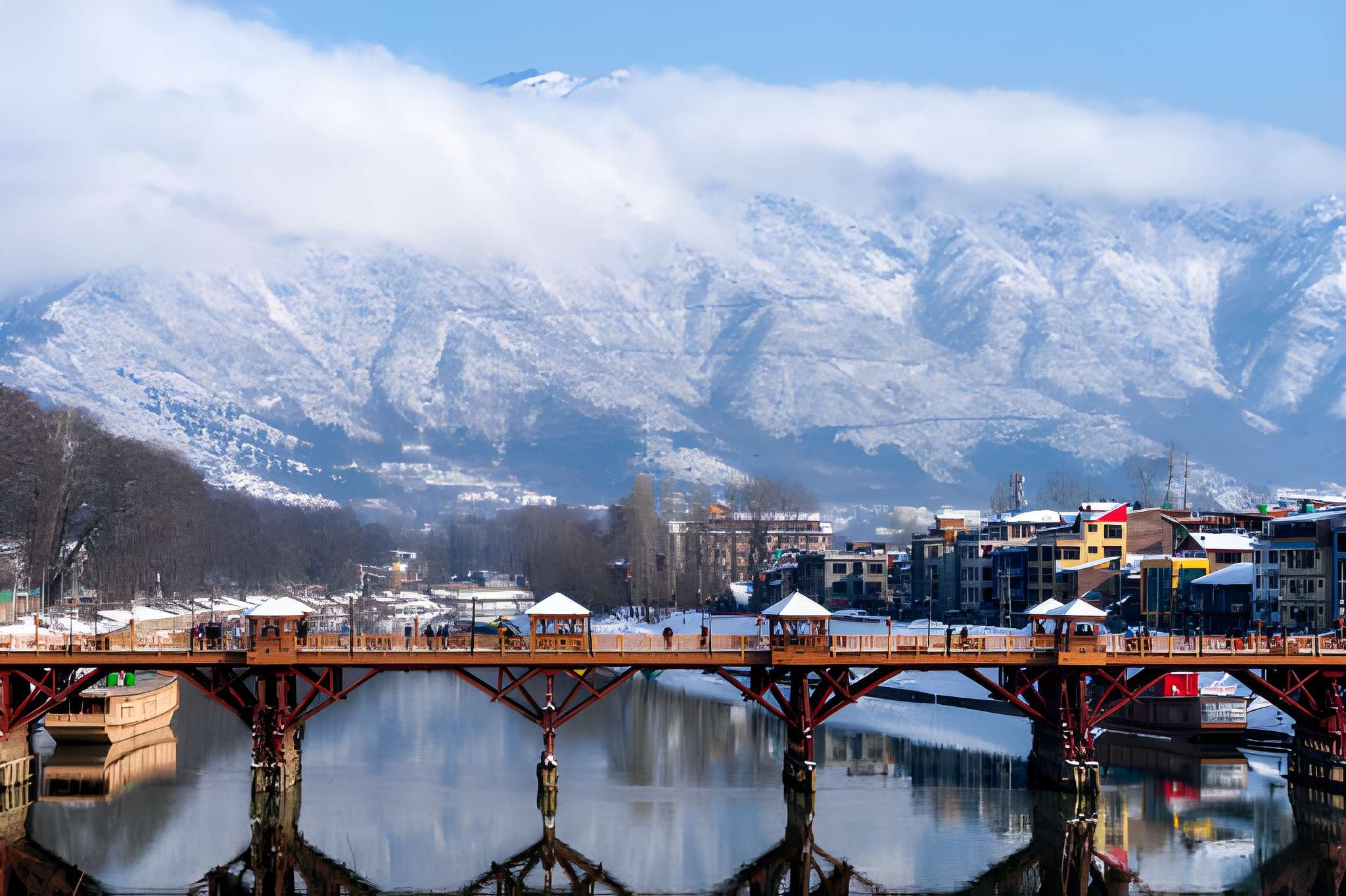 Snow-covered Himalayan town with a wooden bridge over a calm river, buildings along the banks, and clouds drifting above mountain peaks, one of the Top Snowfall Destinations offering a stunning winter landscape in India.