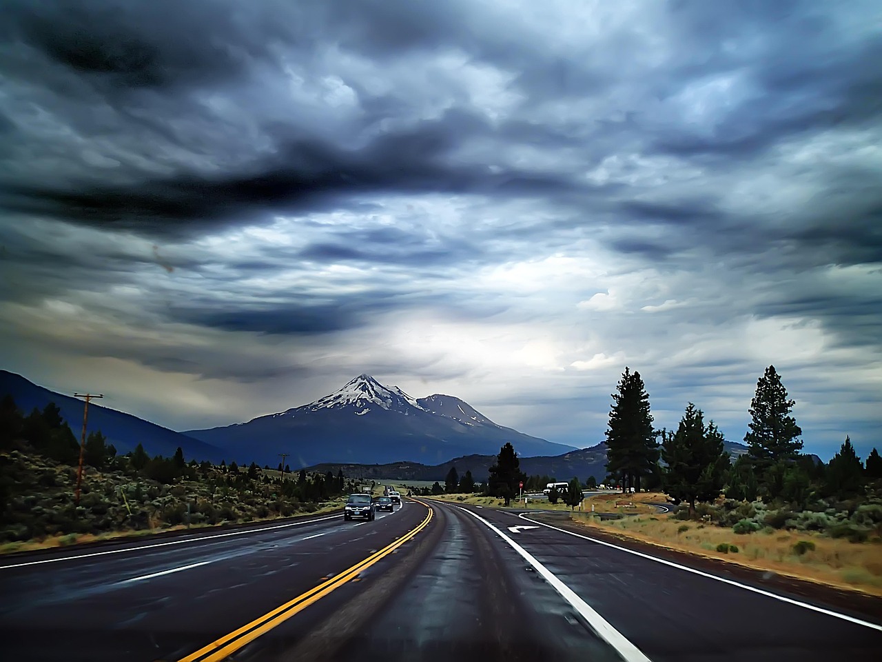 Open highway stretching toward distant snow-capped mountains under heavy dramatic storm clouds, with cars on the road and evergreen trees along the roadside.
