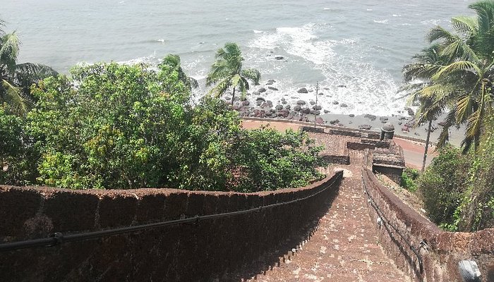 A long stone staircase curving down toward a rocky beach lined with palm trees and crashing waves, offering a scenic coastal experience often enjoyed among the Things to do in Panjim Goa.