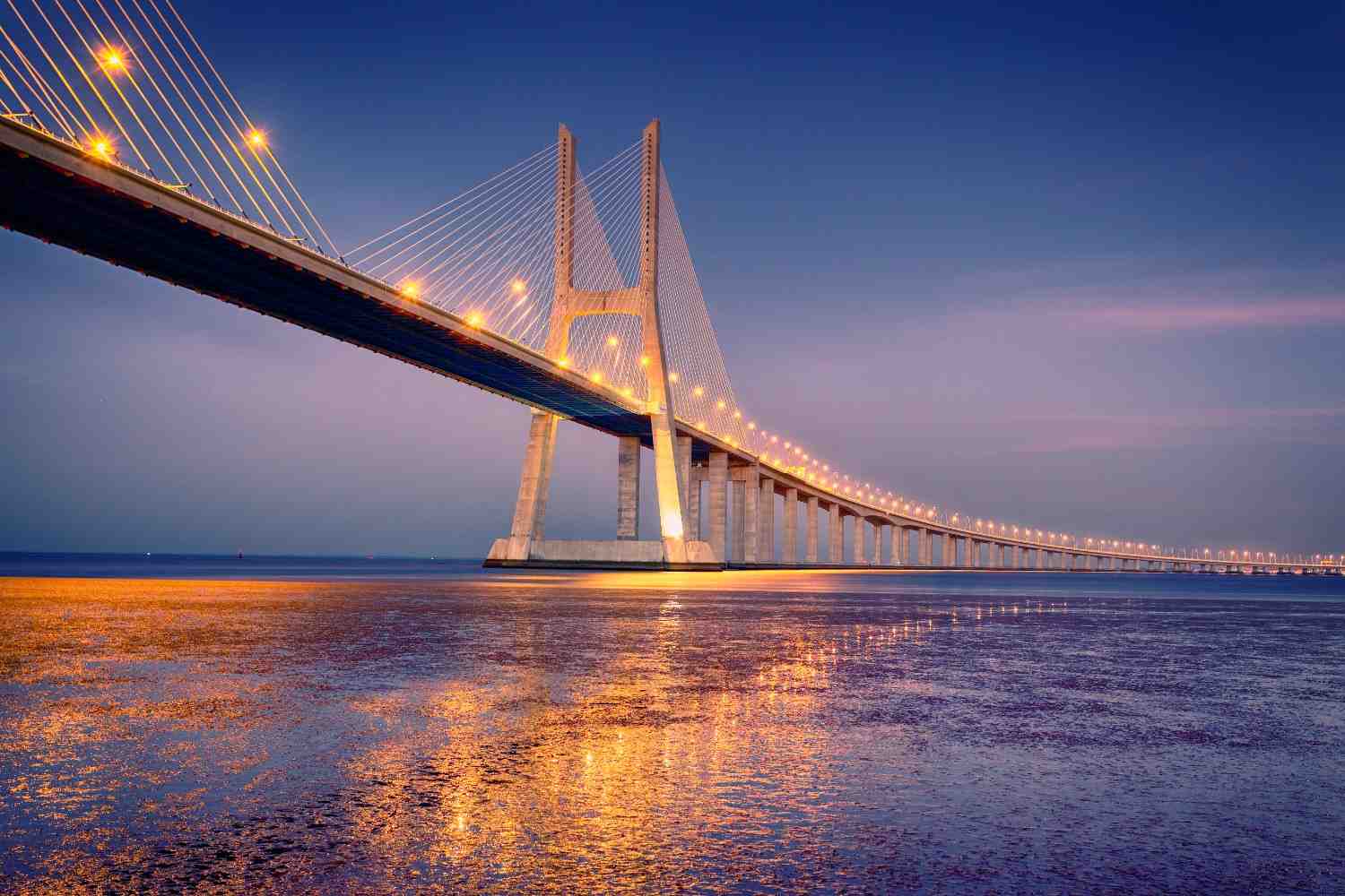 A long illuminated cable-stayed bridge stretching across calm waters during twilight, creating a stunning architectural view that can inspire travelers exploring places to visit in Panjim.
