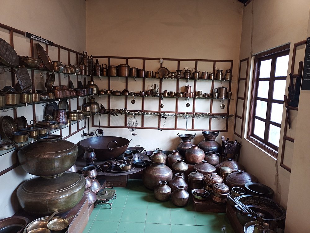 A display room filled with traditional brass and copper utensils, pots, and cookware arranged on shelves and the floor, showcasing heritage kitchen artifacts often seen in Goa museums.