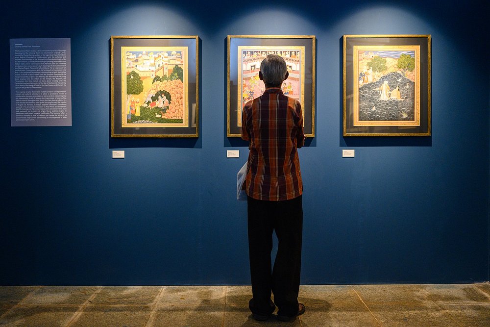 A visitor stands closely observing framed traditional paintings displayed on a deep blue wall, capturing the quiet, immersive atmosphere often found in Art galleries in Goa.