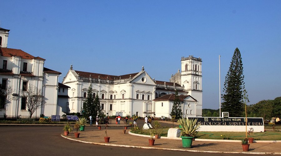 A bright outdoor view of a large white colonial-style building surrounded by gardens and visitors walking along the pathway, part of the Archaeological Survey of India complex often visited by people exploring Museums in Goa.