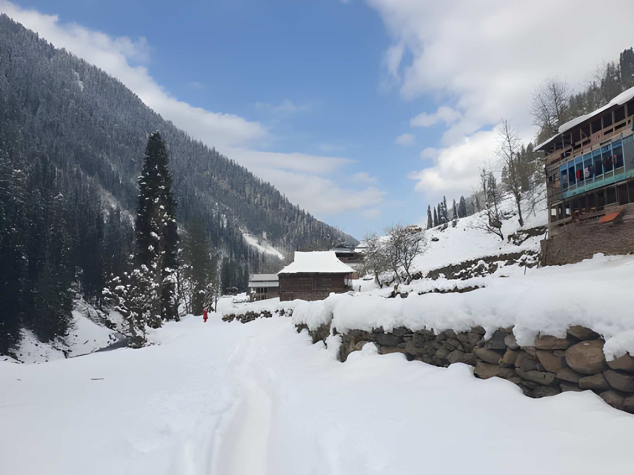 Snow-covered village houses and pine-covered mountains under a clear winter sky, capturing the peaceful charm of December snowfall in India