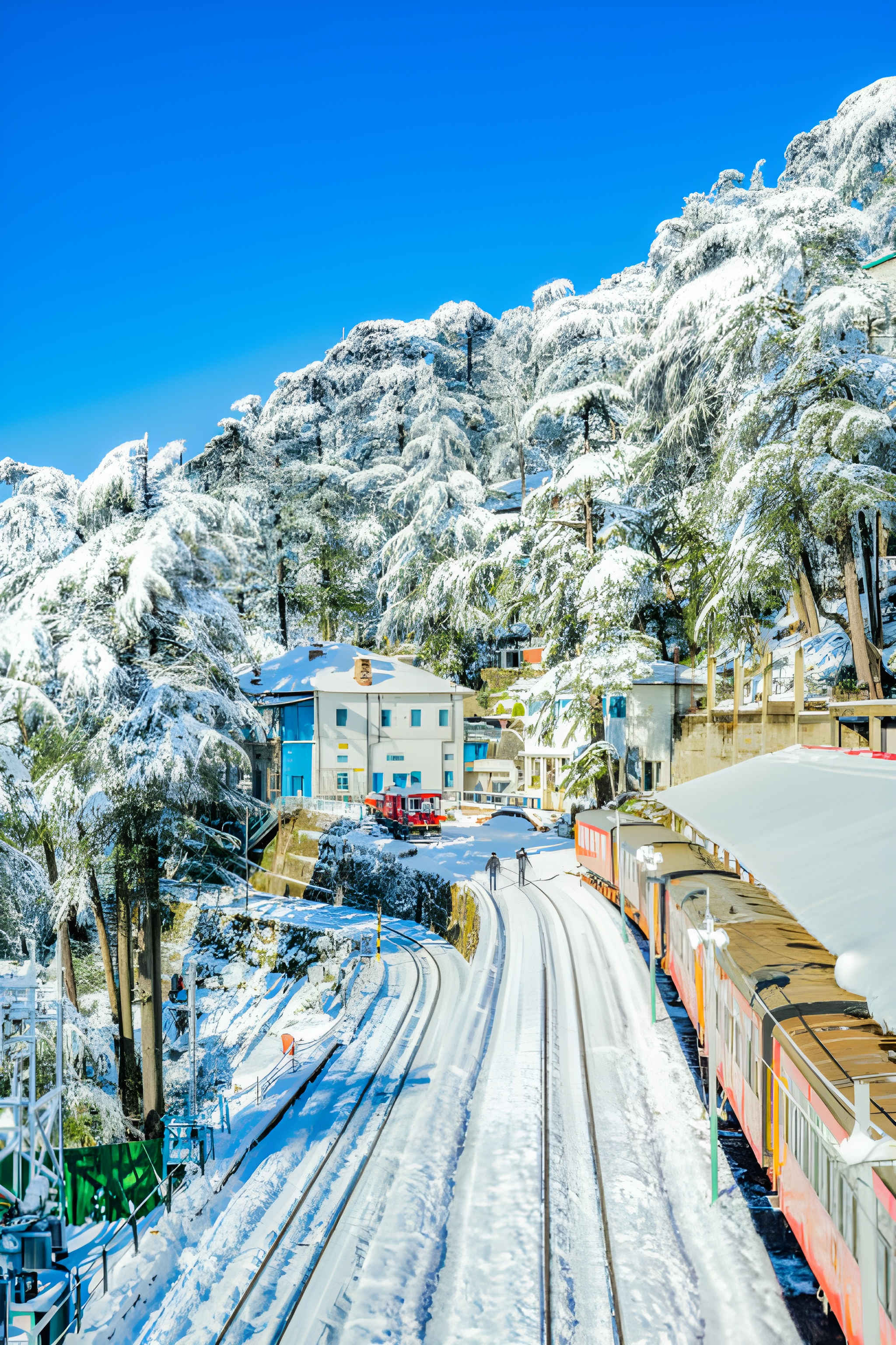 Snow-covered railway tracks, colorful train coaches, and pine trees blanketed in white under a bright blue sky, capturing the charm of snow in India December.